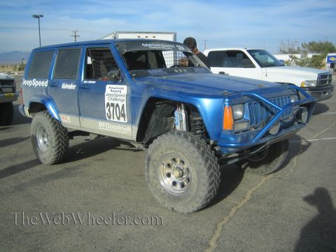 1990 Ford Bronco - blue Jeep Cherokee JeepSpeed race SUV numbered 3704 with a front tube bumper sits in a gravel lot with a box truck behind.