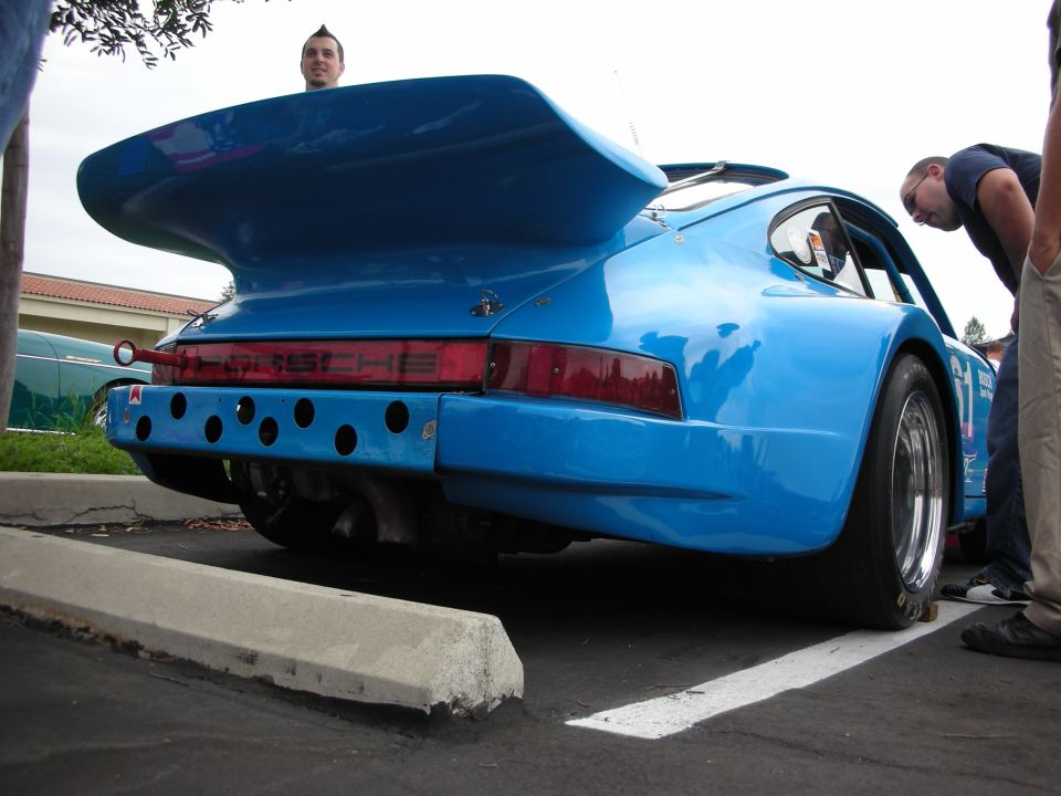 Blue Porsche 911 race car with large rear wing and holes in the bumper. People are looking at it