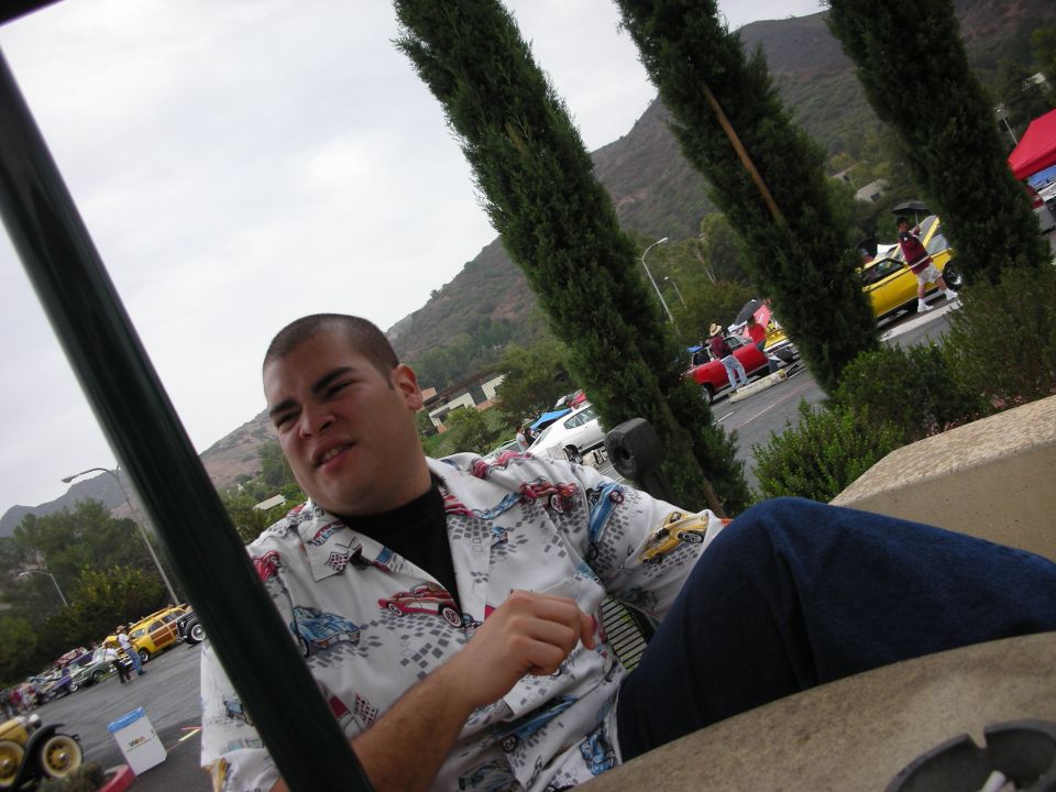 Man wearing a white shirt with car graphics sits in foreground; background shows a car show with several vehicles including a red pickup and a yellow car