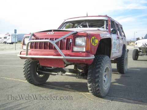 1990 Ford Bronco - red Jeep Cherokee race rig with a tubular front bumper, KC light, lift and oversized tires, photographed from the front on pavement.