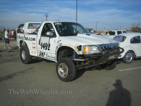 1990 Ford Bronco - white off-road race truck numbered 1492 with SNF Racing and SickNicksFab.com lettering, front bodywork removed, parked at a desert event.