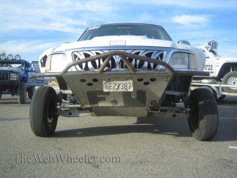 1990 Ford Bronco - Front view of a stripped silver-and-white race truck on a tube front bumper, California plate 6E29387, with other race rigs behind.
