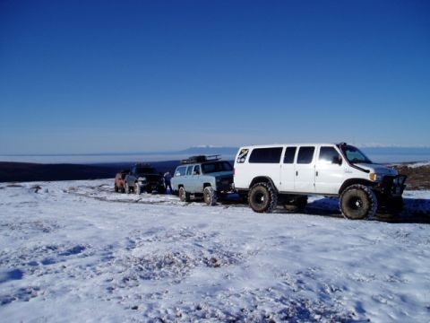2000 Ford E350 - A convoy of modified vehicles, including a white 2000 Ford E350 van, traverses a snowy terrain under a clear blue sky.