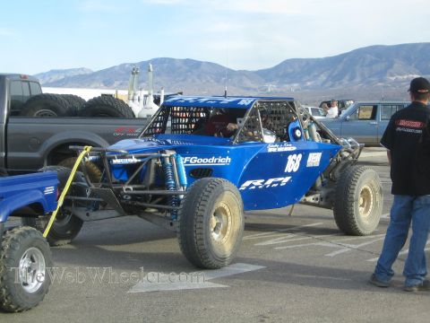 1990 Ford Bronco - blue tube-frame off-road race buggy numbered 168 with BFGoodrich and RPM decals, parked among trucks at a desert staging area.