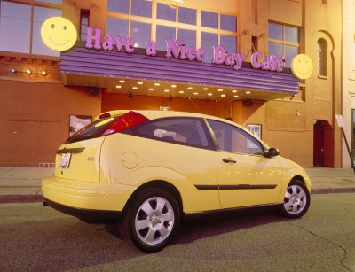 2002 Ford Focus - A yellow 2002 Ford Focus hatchback is parked outside a cafe with a 'Have a Nice Day Cafe' sign.