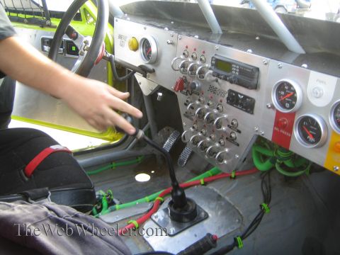 1990 Ford Bronco - Close-up of a race vehicle's aluminum dash panel with rocker switches, a radio and gauges, a hand resting on the shifter beside green wirin…