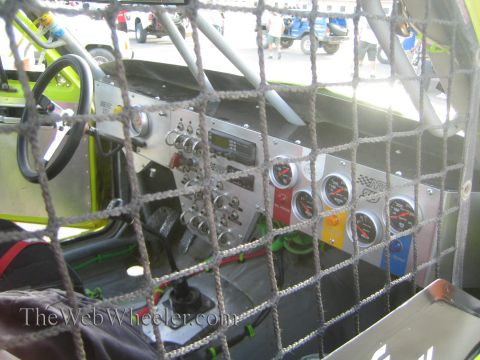 1990 Ford Bronco - Race vehicle interior viewed through a window safety net, showing the aluminum switch panel, gauges and pedals inside the cabin.
