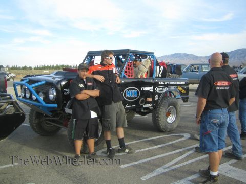 1990 Ford Bronco - pit crew in black team shirts stands beside a black off-road race truck numbered 02 lettered Kal Off Road Racing at a desert event.