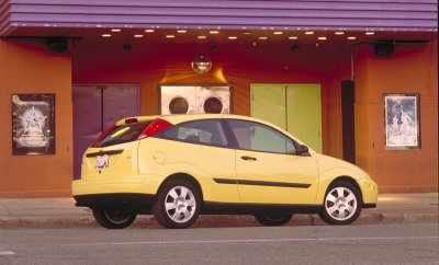 2002 Ford Focus - A yellow 2002 Ford Focus hatchback is parked on the street in front of a building with colorful doors and posters.