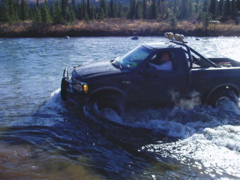 2000 Ford E350 - A dark-colored Ford F-150 truck is shown driving through a body of water, with trees and brush visible on the far bank.
