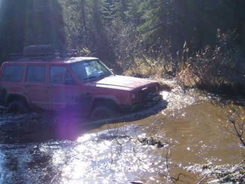 2000 Ford E350 - A red Jeep Cherokee XJ navigates a muddy off-road trail, its front right tire deep in the water.