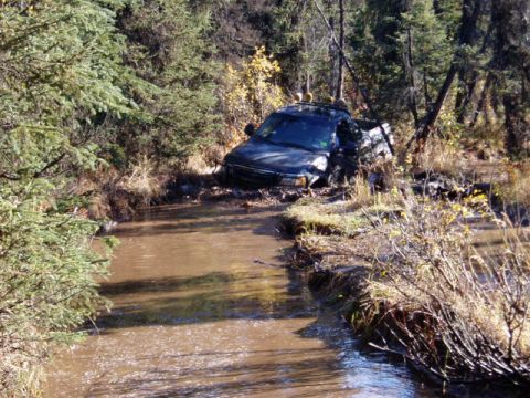 2000 Ford E350 - A dark pickup truck is stuck in a muddy stream, with trees and foliage on either side.