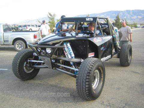 1990 Ford Bronco - black tube-frame off-road race car numbered 129 with BFGoodrich tires, people gathered around it at a desert staging area.