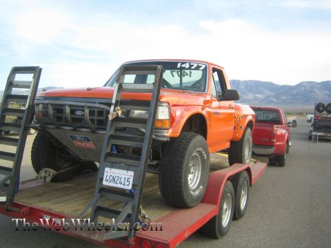 1990 Ford Bronco - orange Ford off-road pickup numbered 1477 strapped on a tilt trailer with loading ramps, mountains in the background.