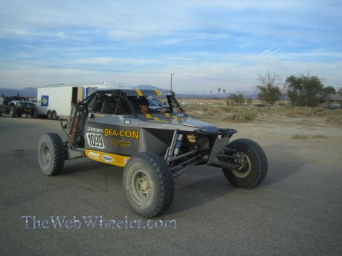1990 Ford Bronco - gray tube-frame off-road race car numbered 1099 with Bea-Con and CAT decals driving on a paved road in the desert.