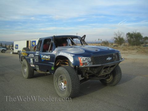 1990 Ford Bronco - blue Chevrolet off-road race truck numbered 159 with Thing Motorsports decals and oversized tires on a paved road in the desert.
