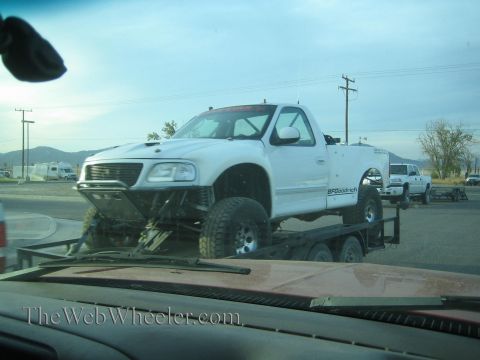 1990 Ford Bronco - white lifted Ford pickup with BFGoodrich tires loaded on a trailer, viewed through the windshield of a following vehicle.