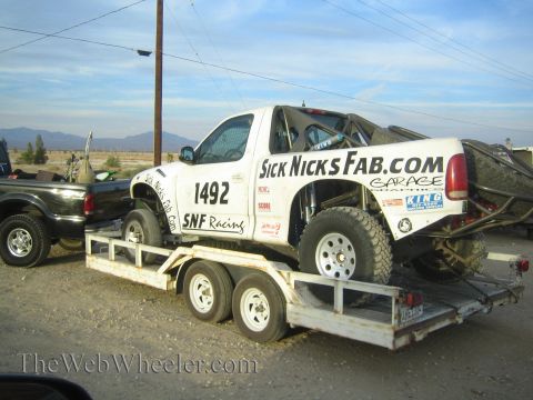 1990 Ford Bronco - white off-road race truck numbered 1492 lettered SickNicksFab.com and SNF Racing, loaded on a tandem-axle trailer in the desert.