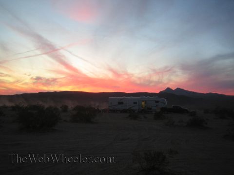 1990 Ford Bronco - pink-and-orange desert sunset over a fifth-wheel RV and truck camped in the dust, with mountains silhouetted on the horizon.