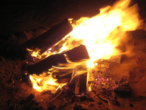 1990 Ford Bronco - nighttime campfire with bright orange flames licking up around split logs over sandy desert ground.