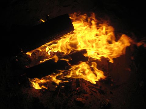 1990 Ford Bronco - Logs burning low in a campfire at night, yellow and orange flames spreading across the dark sand.