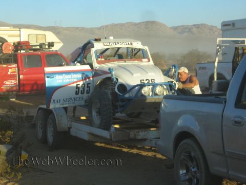 1990 Ford Bronco - white off-road race buggy numbered 535 strapped to a flatbed trailer in a dusty desert pit area, a man standing alongside.