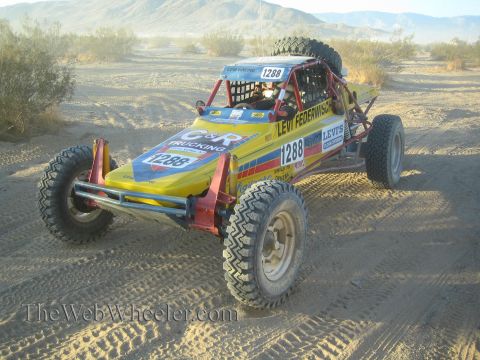 1990 Ford Bronco - yellow and blue open tube-frame off-road race buggy numbered 1288 parked on desert sand with mountains behind.