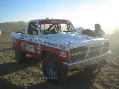 1990 Ford Bronco - red and white off-road race truck numbered 301 with BFGoodrich livery in a dusty desert staging area.