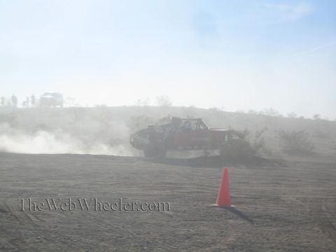 1990 Ford Bronco - off-road race truck throwing up dust as it powers along a desert course past an orange marker cone.