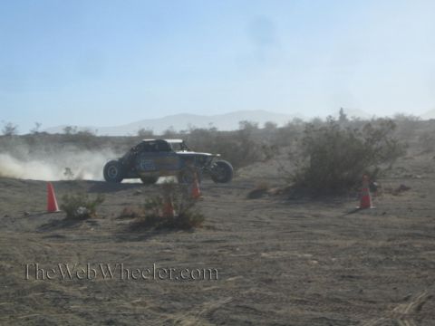 1990 Ford Bronco - off-road race buggy speeding through desert brush, trailing dust between orange marker cones.