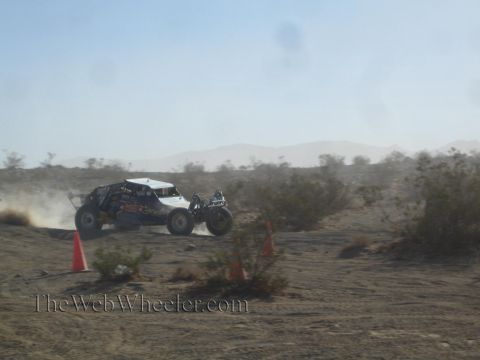 1990 Ford Bronco - open tube-frame off-road buggy racing across dusty desert terrain past orange cones, mountains behind.