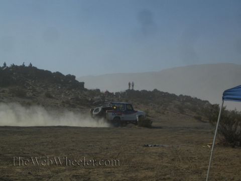 1990 Ford Bronco - off-road race truck kicking up dust as it rounds a desert hill, with spectators silhouetted on the ridge.