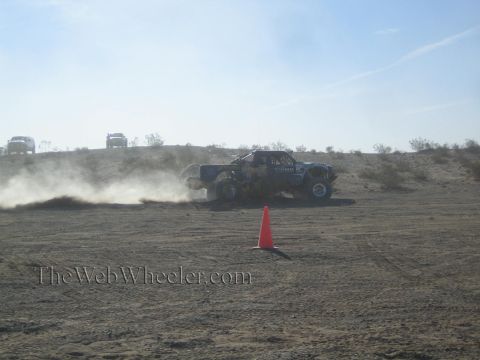 1990 Ford Bronco - blue off-road race truck sliding through the desert in a cloud of dust beside an orange marker cone.