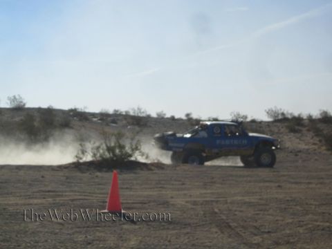 1990 Ford Bronco - blue off-road race truck racing along a dusty desert course, throwing dust beside an orange cone.