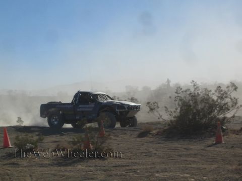 1990 Ford Bronco - black off-road race truck cornering hard in a haze of dust between orange marker cones in the desert.