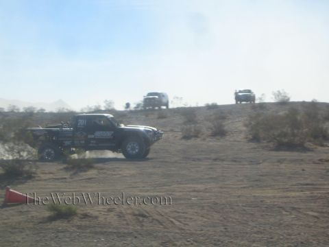 1990 Ford Bronco - black off-road race truck numbered 701 racing across the desert with support trucks visible on the ridge behind.