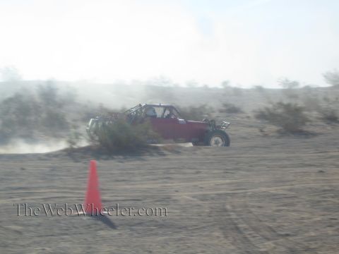 1990 Ford Bronco - red open-frame off-road buggy speeding through a dusty desert course past an orange marker cone.
