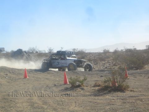 1990 Ford Bronco - white tube-frame off-road buggy racing across dusty desert ground between rows of orange cones.
