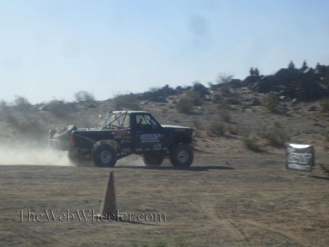 1990 Ford Bronco - dark Ford off-road race truck cornering in a cloud of dust, spectators watching from the desert ridge.