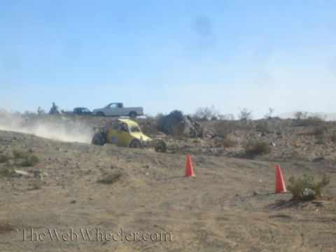 1990 Ford Bronco - yellow off-road buggy racing through desert brush, a white pickup parked on the rise behind it.