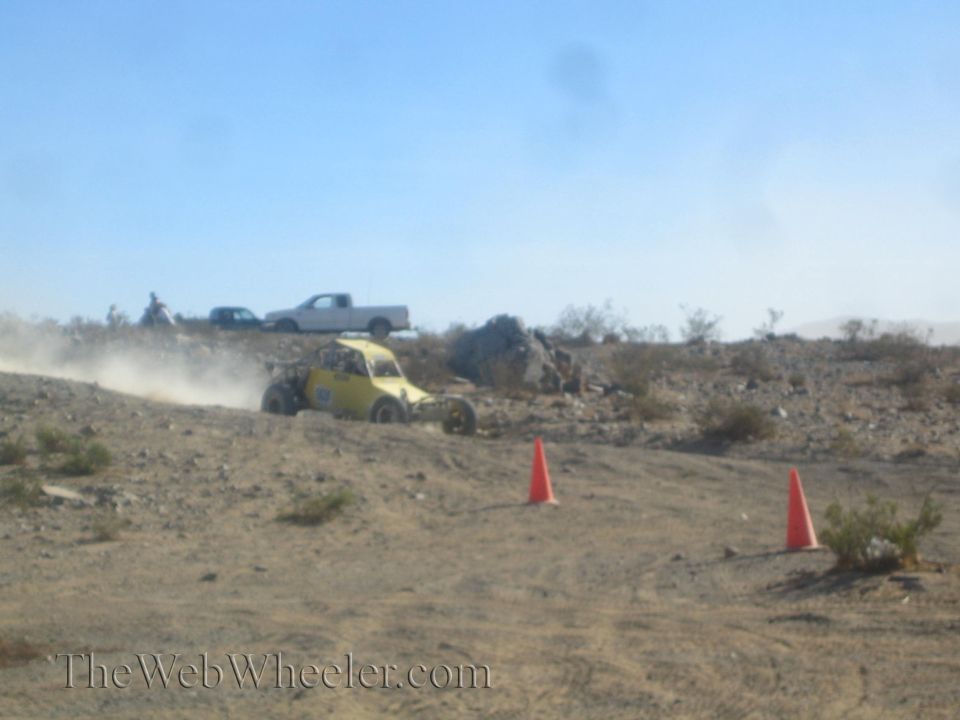 Yellow off-road buggy racing through desert brush with a pickup parked behind