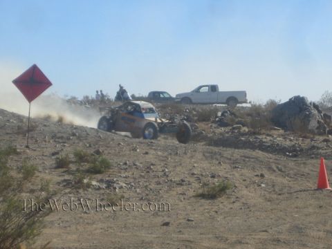 1990 Ford Bronco - off-road race buggy speeding through the desert past a red marker flag, with trucks and spectators behind.