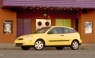 2002 Ford Focus - A yellow 2002 Ford Focus hatchback is parked on the street in front of a building with colorful doors.