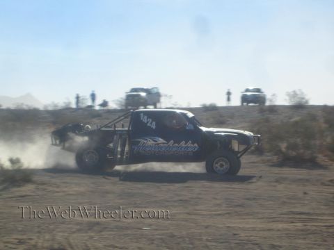 1990 Ford Bronco - black off-road race truck numbered 1A24 racing across the desert trailing dust, chase trucks on the ridge.