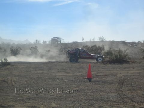 1990 Ford Bronco - red tube-chassis off-road race truck speeds across a dusty desert course past an orange marker cone.