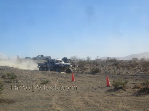 1990 Ford Bronco - blue off-road race truck throws up dust on a rocky desert track marked by two orange cones, with spectator trucks behind.