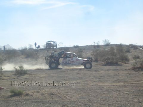 1990 Ford Bronco - white and red sponsor-decaled off-road race buggy races across the desert trailing a plume of dust.