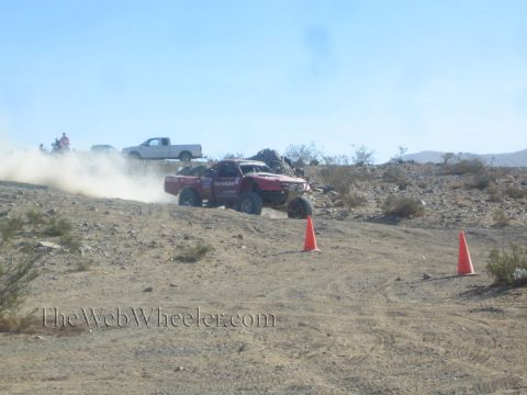 1990 Ford Bronco - red off-road race truck crests a sandy desert rise between orange marker cones as dust trails behind.