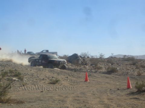 1990 Ford Bronco - black off-road race truck speeds across rocky desert terrain, kicking up dust near two orange cones.