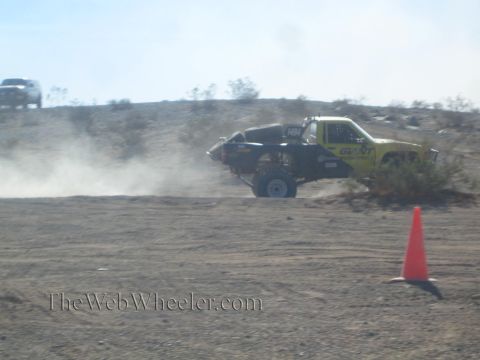 1990 Ford Bronco - yellow and black off-road race truck catches air over a desert berm, trailing dust past an orange cone.